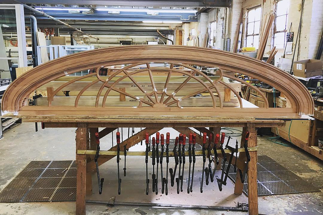 A curved fanlight under construction in the Avenel workshop: a mahogany arch with radiating muntins and a carved centre, clamped along its base on the bench